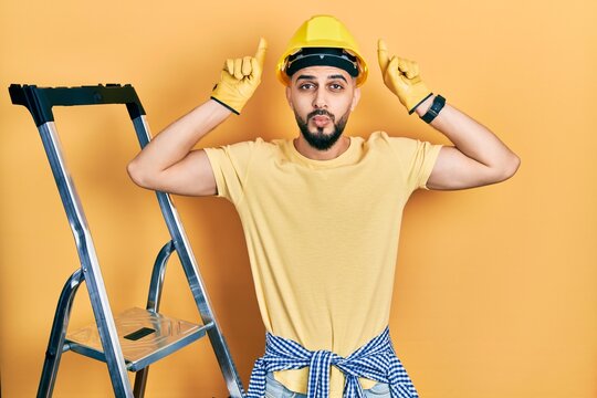 Handsome man with beard by construction stairs wearing hardhat doing funny gesture with finger over head as bull horns