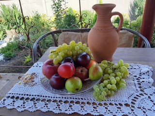 In the garden, on the table, on a white lace napkin, there is a jug and a plate with fruits: apples, pears, plums, grapes.