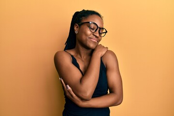 African american woman with braided hair wearing casual clothes and glasses hugging oneself happy...