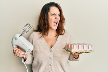 Young caucasian woman holding pastry blender electric mixer and eggs angry and mad screaming...