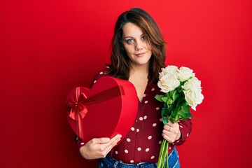 Young caucasian woman holding valentine gift and flowers relaxed with serious expression on face. simple and natural looking at the camera.