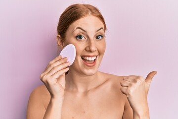 Young irish woman holding makeup sponge pointing thumb up to the side smiling happy with open mouth