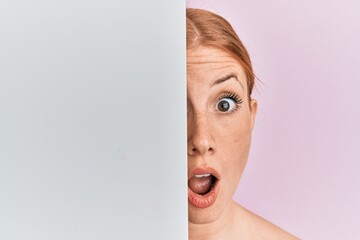 Young irish woman holding blank empty banner showing half face afraid and shocked with surprise and...