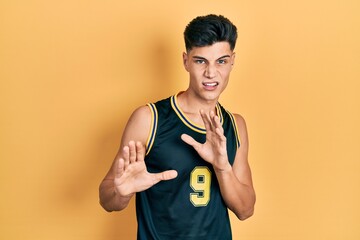 Young hispanic man wearing basketball uniform disgusted expression, displeased and fearful doing...