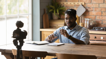 Happy Afro American blogger guy taking video on dslr camera fixed on tripod, shooting post at home for vlog. Virtual coach, influencer recording lecture, filming training speech for online channel