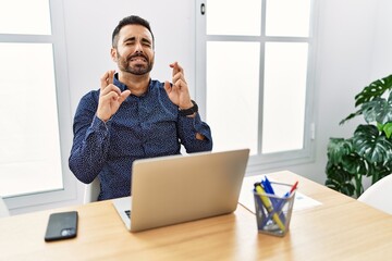 Young hispanic man with beard working at the office with laptop gesturing finger crossed smiling with hope and eyes closed. luck and superstitious concept.