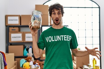 Young hispanic man wearing volunteer t shirt holding money from charity jar scared and amazed with...