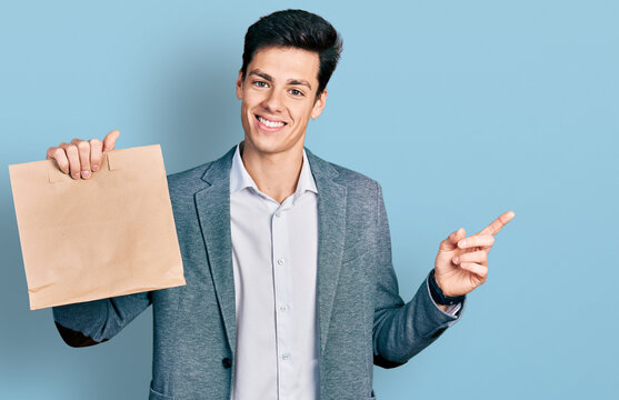 Young hispanic business man holding paper bag with bread smiling happy pointing with hand and finger to the side