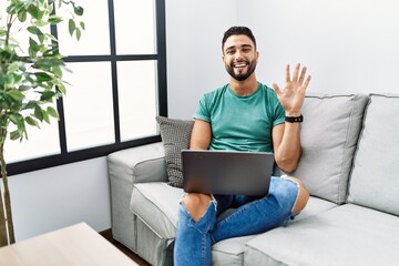 Young handsome man with beard using computer laptop sitting on the sofa at home showing and...