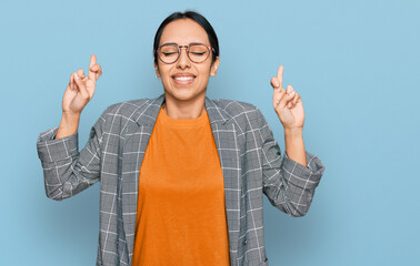 Young hispanic girl wearing business jacket and glasses gesturing finger crossed smiling with hope and eyes closed. luck and superstitious concept.
