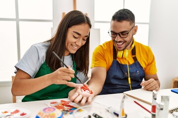 Fototapeta premium Young hispanic couple smiling happy painting hands sitting on the table at art studio.