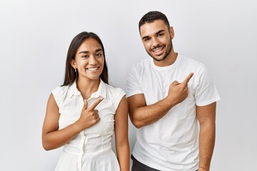 Young interracial couple standing together in love over isolated background cheerful with a smile of face pointing with hand and finger up to the side with happy and natural expression on face