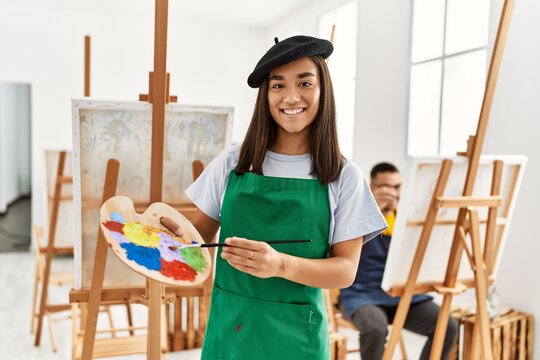 Young hispanic couple drawing at art studio. Woman smiling happy and holding paintbrush and palette.