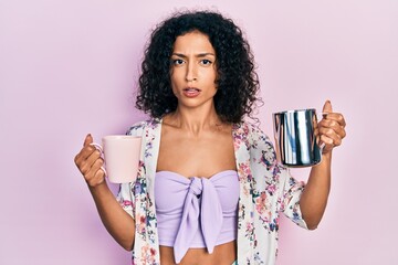 Young latin girl holding cup of coffee and jar of milk clueless and confused expression. doubt...