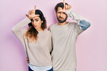 Young hispanic couple wearing casual clothes making fun of people with fingers on forehead doing...