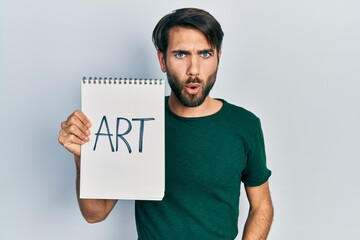 Young hispanic man holding art notebook scared and amazed with open mouth for surprise, disbelief...