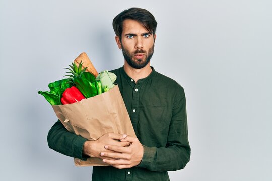 Young hispanic man holding paper bag with bread and groceries scared and amazed with open mouth for surprise, disbelief face