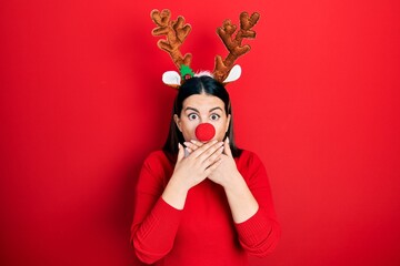 Young hispanic woman wearing deer christmas hat and red nose shocked covering mouth with hands for mistake. secret concept.