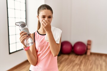 Young brunette teenager wearing sportswear holding water bottle smelling something stinky and...