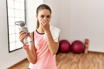 Foto op Canvas Young brunette teenager wearing sportswear holding water bottle smelling something stinky and disgusting, intolerable smell, holding breath with fingers on nose. bad smell © Krakenimages.com