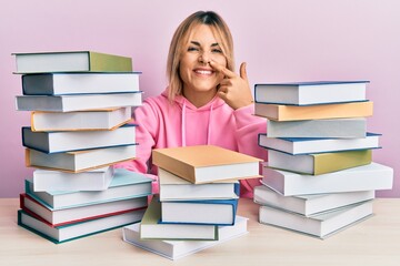 Young caucasian woman sitting on the table with books pointing with hand finger to face and nose, smiling cheerful. beauty concept