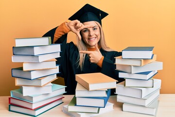 Young caucasian woman wearing graduation ceremony robe sitting on the table smiling making frame with hands and fingers with happy face. creativity and photography concept.