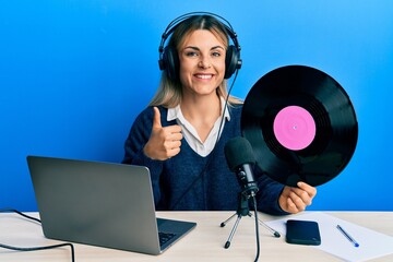 Young caucasian woman working at radio studio holding vinyl disc smiling happy and positive, thumb up doing excellent and approval sign