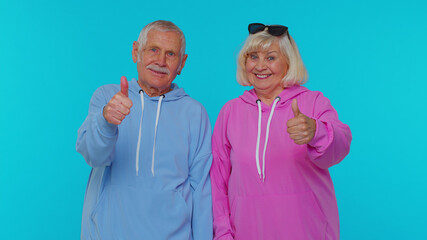 Senior stylish grandparents pensioners looking approvingly at camera showing thumbs up, like sign positive something good isolated on blue background. Elderly mature retired grandfather grandmother