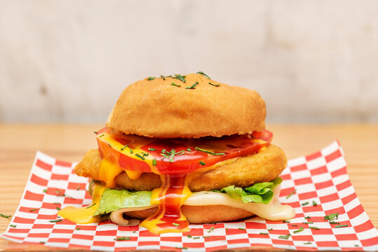 Close-up image of breaded chicken burger with fresh lettuce, tomato slice and mustard sauce with ketchup dripping on melted cheese and red and white squared paper