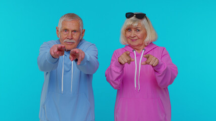 We choose you. Elderly woman granny with man grandfather pointing to camera looking with happy expression, making choice, showing direction. Senior grandmother with grandpa isolated on blue background
