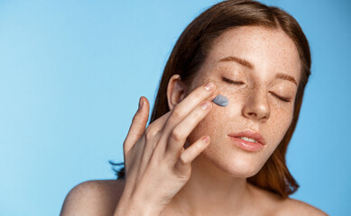 Beauty and spa. Redhead girl applies clay mask, scrub or facial cream on face, enjoyes softness of...