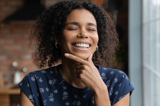 Portrait of happy beautiful millennial African woman touching chin, facial skin with toothy smile and closed eyes, enjoying beauty care effect, standing in home interior. Young Black girl headshot