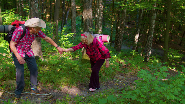 Senior grandmother grandfather walking, enjoying hikinig adventures. Mature woman holding mans hand helping her climb on mountain hill. Elderly grandparents tourists traveling with backpacks in wood