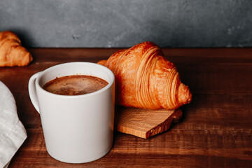 Fresh baked croissants, mug of black coffee on brown wooden table. Cup of hot Americano with foam,...