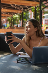 beautiful young asian woman in pink dress with dark long hair drinking coffee. attractive smiling student lady having rest on veranda of cafe with laptop. summer chill, rest