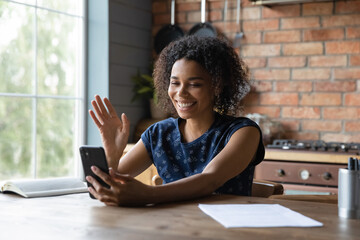 Happy millennial African mixed race girl using smartphone, making video call from home, waving hello, looking and speaking at webcam, smiling at camera, talking on cell, attending virtual conference