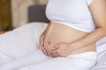 Fototapeta na wymiar Close up Happy Pregnant Woman sitting on bed holding and stroking her big belly at home,Pregnancy of young woman enjoying with future life relax at home,Motherhood and Pregnant Concept