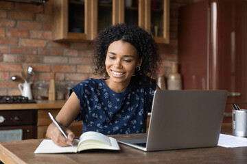 Happy smiling mixed race African student girl studying from home, writing notes, watching, listening, summarizing learning webinar on laptop, attending virtual school class, online college lesson
