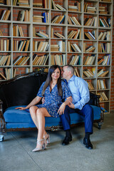 Obraz premium stylish man in a blue shirt with a beautiful wife in a dress are sitting together on a cozy sofa against the background of a luxurious room with books. Family happiness