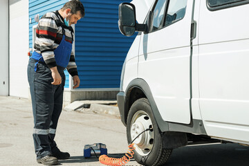A mechanic driver in overalls pumps the wheel of a truck with a hydraulic pump on the street on a summer day. A real scene of a wheel breakdown or puncture on the road.