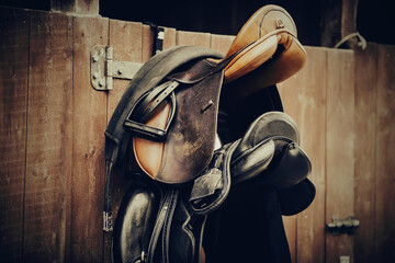 Brown and black saddles hang on the stall. Horse ammunition. - obrazy, fototapety, plakaty