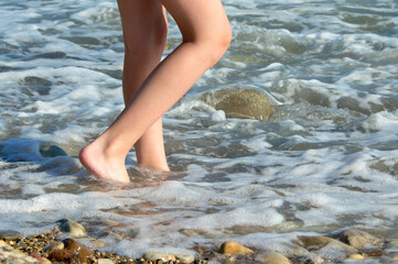 The girl's legs go into the sea surf on a pebble beach in the sun. The concept of tourism.