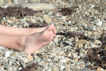 The girl's legs are wrinkled from the water on a pebble beach in the sun. The concept of tourism.