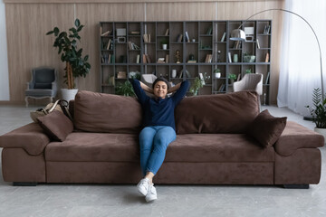 Happy calm young Indian woman sit relax on couch in design living room take nap or sleep. Peaceful...
