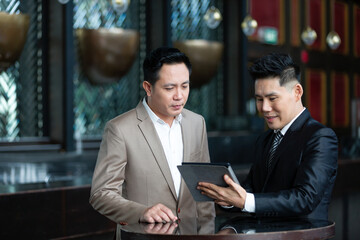 Group of Asian businessman wearing suits talking using digital tablet together while standing in...