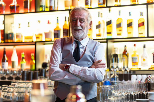 Smiling senior barman or waiter in apron on wine or alcohol blur background. Senior bartender smiling with arms crossed at the bar. alcohol, people, service concept
