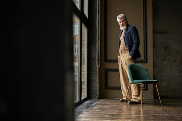 Full length shot of classy grey haired middle aged man in casual wear looking at camera while standing by a large window in modern loft interior