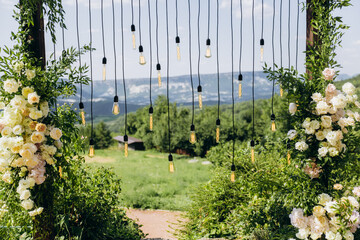 Fototapeta premium Wedding arch on rustuc style with white flowers in park with light bulbs.