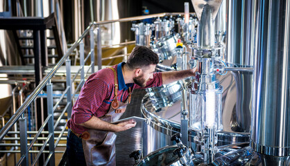 Young male brewer in leather apron supervising the process of beer fermentation at modern brewery factory