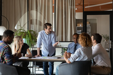 Confident young Caucasian businessman hold lead meeting with diverse multiethnic employees colleagues in office. Motivated male leader or boss talk at team negotiation briefing. Leadership concept.